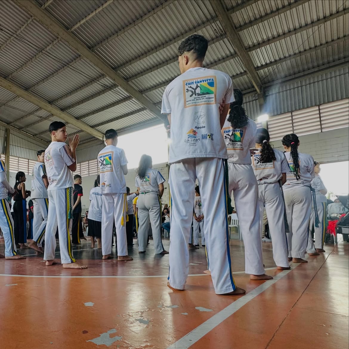 Capoeiristas em roda de pé, de uniforme branco, dentro de ginásio